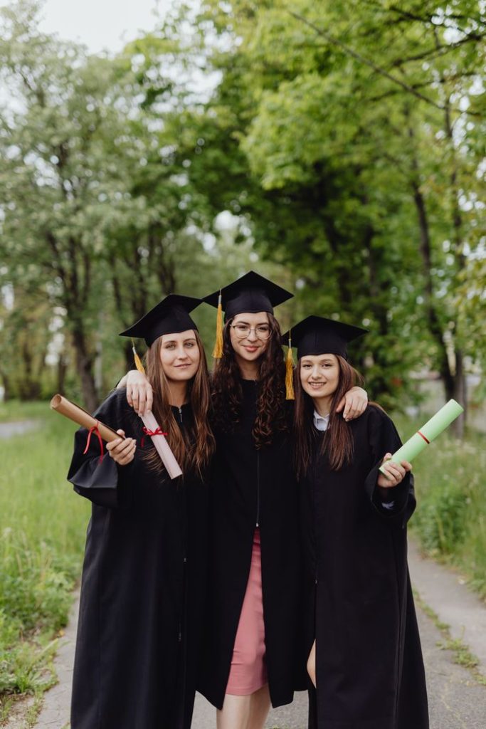 Three young women in graduation gowns celebrating together outdoors.